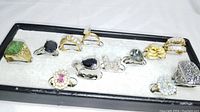 Twelve assorted vintage fashion rings in mixed metals displayed in a single row on a white padded tray with black frame. Variety of shapes and stones, including green, black, pink, and clear stones as well as floral and filigree designs.