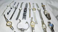 Collection of 15 women's wristwatches arranged on white cloth, showing various styles, metals, and dial colors.
