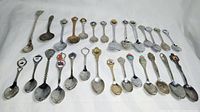 Front angled view of 28 different collectible miniature state spoons arranged in four rows on a white fabric background showing various state emblems and decorative handles.