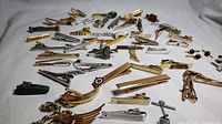 Photo showing a wide assortment of vintage tie clips and alligator pins laid out on a white surface.