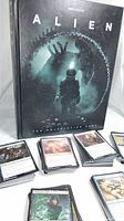 Photo showing Alien Free League RPG book standing upright with stacks of Magic The Gathering cards spread out in front.