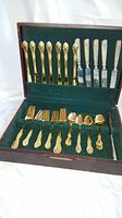 Wide view of the open wooden storage case containing the gold flatware set (forks, spoons, and knives) and silver-toned cutting tools.
