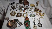 Overview of an assortment of metal trinkets including US army pins, miniature picture frames, keychains, whistles, bottle openers, belt buckles, a tea strainer, small chair, and car figurines.