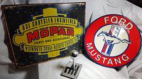 Photo showing the Mopar rectangular metal sign, Ford Mustang round metal sign, and the silver shifter knob paperweight together on a white fabric background.