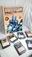 Photo of 'TRIAX and the NGR' softcover RPG book with several stacks of Magic The Gathering cards arranged in front, showing card fronts.