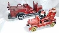 Two cast iron fire trucks shown from side angle, displaying firefighter figure and hose reel on smaller truck and chassis of larger truck