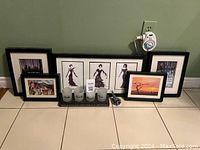 Photo shows seven framed art prints in black frames arranged on floor against wall, four frosted candle holders with 'Yankee Candle' branding in front, and a small metallic item. Power outlet and plug-in device visible on wall.