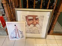 Two framed art prints leaning against wooden stair railing. The larger print is a Santa Claus portrait art print described as light-up art. The smaller print depicts a woman in a pastel pink dress with a large bow on her back.