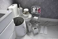 Photo showing a variety of white and chrome bathroom accessories including trash bin, tissue box holder, toilet brush holder, and multi-tier wire shelf unit.