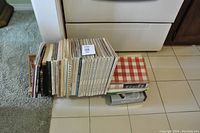 Photo showing the assortment of cook books stacked on the floor, next to a kitchen stove.