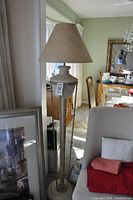 Tall floor lamp with beige fabric shade beside a beige chair with linens draped on the seat, dining room visible in background.
