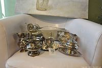 Photo of assorted plated silverware pieces arranged on a white chair, showing pitchers, bowls, and a covered dish.