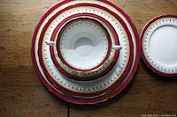 Top view of stacked Aynsley china dinnerware showing the red borders with gold details, includes cup, saucer and plate.