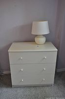 Photo of a white bedside table with three drawers and a small white lamp with round ceramic base and white shade on top.