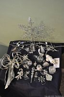 Various silver Christmas ornaments arranged on a black cloth including bows, snowflakes, birds and more.