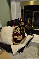 Photo of teddy bear, sherpa blanket, plaid pillow, and two standing Christmas dolls arranged in front of a fireplace.