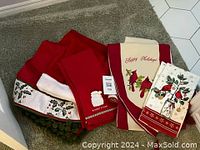 Photo of Christmas-themed linens and a red holiday shirt laid on a rug, showing various embroidered holiday designs such as poinsettias, holly, birds, and festive text.