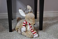 Straw reindeer with red and white striped scarf and white pom-poms positioned on carpet.
