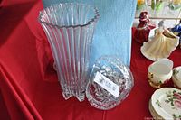 Photo of clear crystal vase with vertical ridges and scalloped rim next to a clear cut crystal bowl with geometric cut patterns, placed on a table with other decorative items in background.