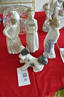 Five Lladro porcelain figurines on a red cloth background: three children, one girl holding books, one boy holding blue cloth, one child wearing a hat, and a small black and white dog figurine.