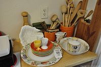 Photo of assorted dishware, colorful plates, egg cups, and wooden kitchen utensils on a wooden shelf.
