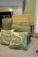 Four outdoor decorative pillows, two solid green and two with green leaf pattern, placed in front of two wooden TV dinner stands.
