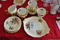 Photo showing 3 teacups stacked inside one another, 3 matching saucers stacked, 7 small plates stacked, creamer, and square cake platter with floral pink rose pattern and gold trim, arranged on red cloth surface.