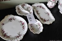 Assorted serving plates, cake plate, and serving trays with floral pattern and gold scalloped edges on black background.