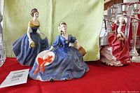 Photo of two Royal Doulton ladies figurines Elyse and Melanie wearing blue dresses, displayed on a red cloth with a green background, showing glossy glaze and detailed sculpting.