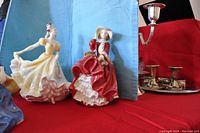 Two Royal Doulton figurines, one of a lady in a yellow dress and one of a lady in a red dress, displayed on a red cloth with a blue backdrop and silver candlesticks nearby.
