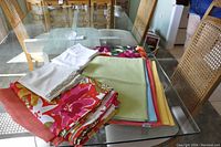 Photo showing assorted folded cloth placemats and tablecloths in various colors and floral patterns on glass table.