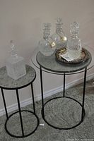 Two round side tables with mosaic tops and metal frames, holding three crystal decanters, two round on metal tray and one square on smaller table.