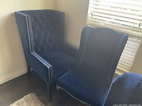 Pair of blue velvet upholstered wingback accent chairs with button tufting and silver nailhead trim on wooden legs.