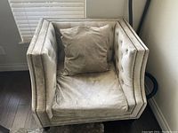 Beige fabric armchair positioned against a wall in a room with a window and hardwood flooring.
