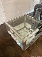 Square mirrored glass coffee table placed on wooden floor next to a white wall and metal chair.