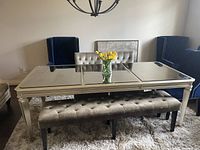 Dining table with mirrored top, tufted bench underneath, two gray tufted chairs behind, and two blue wingback chairs at table ends in carpeted room with wall and light fixture visible