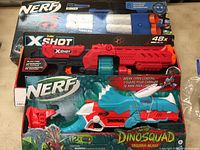Photo showing three boxed toy blasters: Nerf Roblox Arsenal Pulse Laser, Nerf Dinosaurs Tricera-Blast, and Zuru X-Shot blaster.