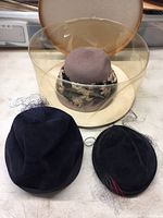 Three assorted vintage hats including beige wide-brim, taupe pillbox with lace, and black velvet pillbox with veil and feather.