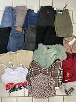Overview of folded pants in blue, gray, black, olive green colors, assorted tops including beige sweater, white floral bralette, green fuzzy jacket, plaid and heart-patterned blouses, and red Nike USC hoodie.