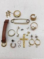 Top-down image showing various vintage jewelry pieces including rings, earrings, a gold cross pendant, small hoops, a pearl brooch, and a brown leather strap.