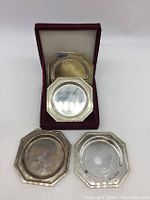 Four octagonal silver-tone coasters displayed with a red carrying case inside which two coasters are placed.
