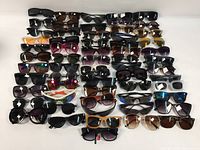 Overhead image showing approximately 50 assorted pairs of sunglasses with various frame styles, shapes, and lens colors arranged in rows on a white background.