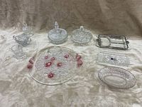 Nine crystal and glass pieces displayed on cream fabric: two covered dishes with lids, round platter with red flowers, compartmentalized tray, crystal design trays, small circular platter, cream sugar bowl, small basket with handles.