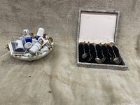 View of six silver spoons neatly arranged in velvet-lined presentation box with leopard print exterior. Next to the box is a decorative white dish containing a variety of souvenir thimbles in different colors and designs.
