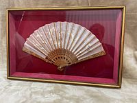 Rectangular frame with glass front showing a mounted cream colored Chinese fan with gold and black painted details and cut-out pattern on wooden base sticks, mounted on deep red fabric background.