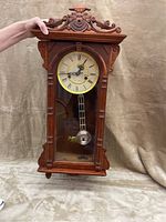 Front view of a vintage grandmother clock with ornate carved wooden frame and glass door showing pendulum and clock face