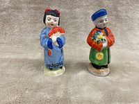 Front view of two Japanese ceramic salt and pepper shakers, female figure with fan and male figure with floral bouquet, standing on beige cloth.