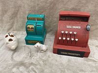 Photo showing the red Tom Thumb cash register, turquoise Nickel's Dime Quarter cash register, and two piggy banks, a cow and a small white animal, displayed on a beige fabric.
