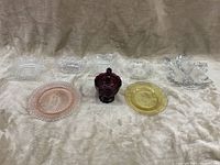 Nine glass and crystal pieces arranged on a beige fabric. Seven clear crystal dishes with various shapes and intricate patterns, one deep amethyst covered jar in center, one pink depression glass plate, and a small yellow glass plate.