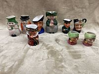 Overview of nine various Toby mugs arranged on a beige fabric surface showing their detailed character features and colorful paint.